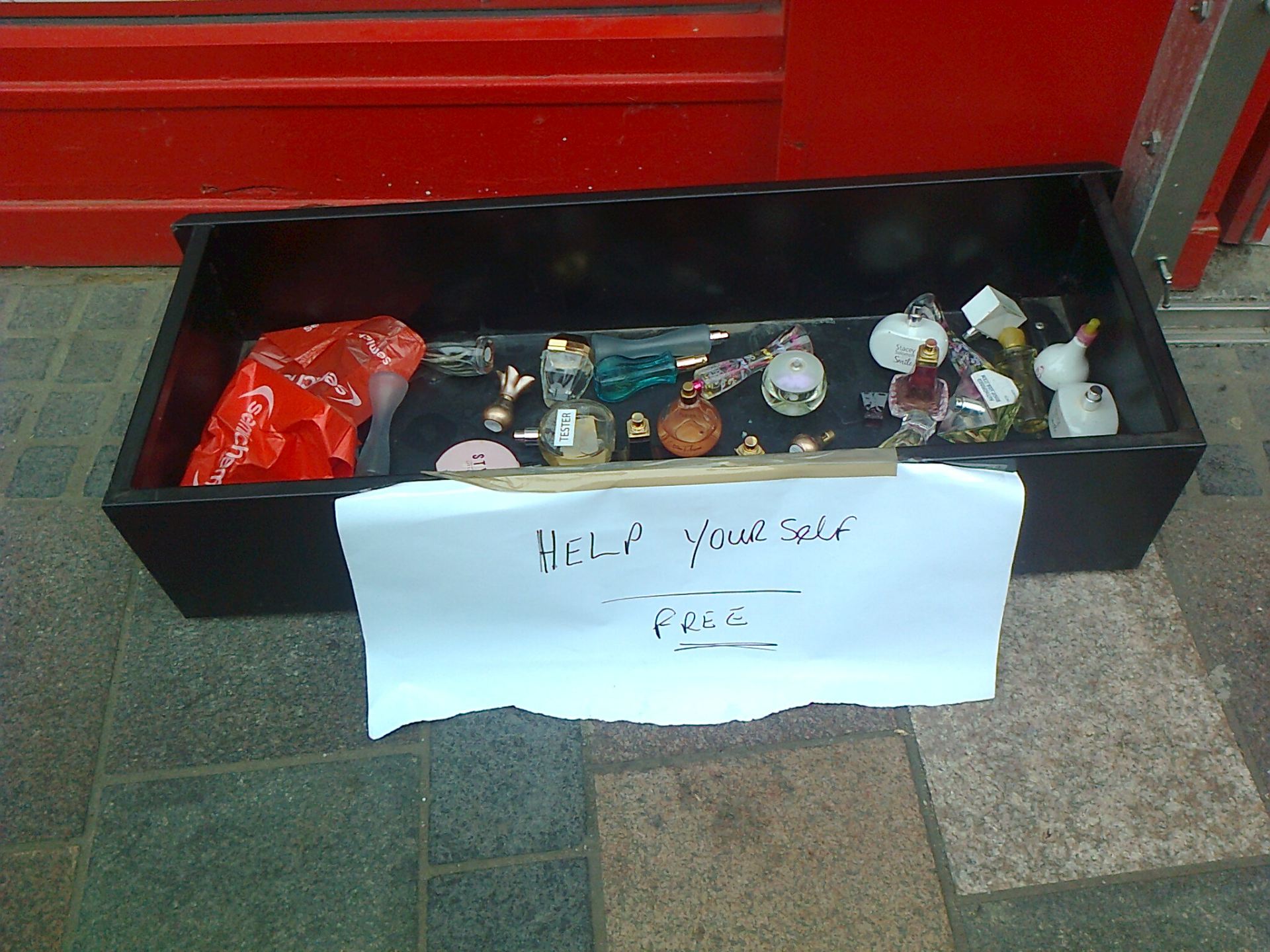 A box of perfume testers lies on a pavement with a sign telling people to help themselves for free.