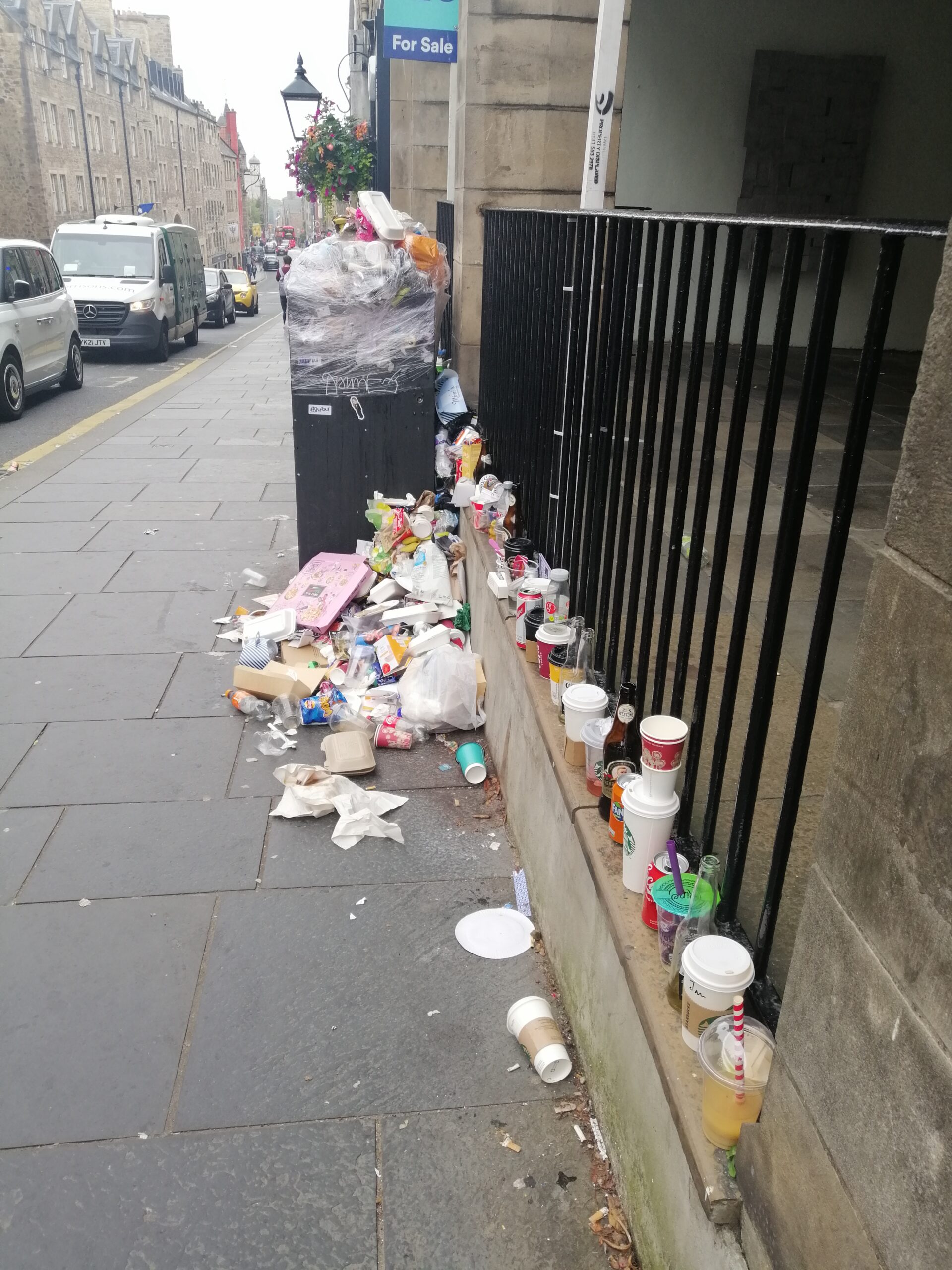 An overflowing bin on the Royal Mile has been wrapped in clingfilm. It is surrounded by take-away containers, cups, bottles, cans and other waste.