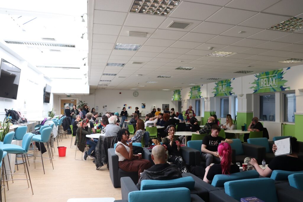 People sitting on comfortable chairs and around tables, with some people standing in front of the far wall.