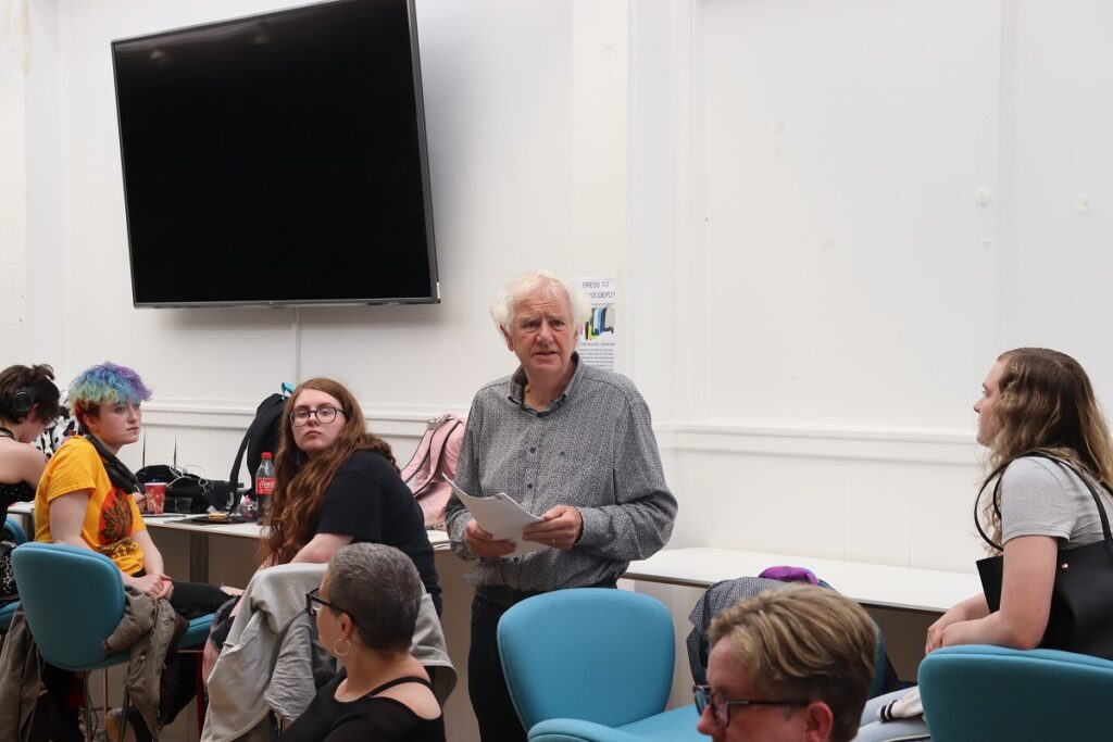 A man in a grey shirt reads from a piece of paper, while several audience members look on.