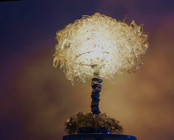 A photograph of a lamp that has been constructed out of waste plastic. The bulb is surrounded by scraps of clear plastic, from bottles. The lamp stand is made from plastic bulb bases. The whole thing looks like a brain on a spinal cord.